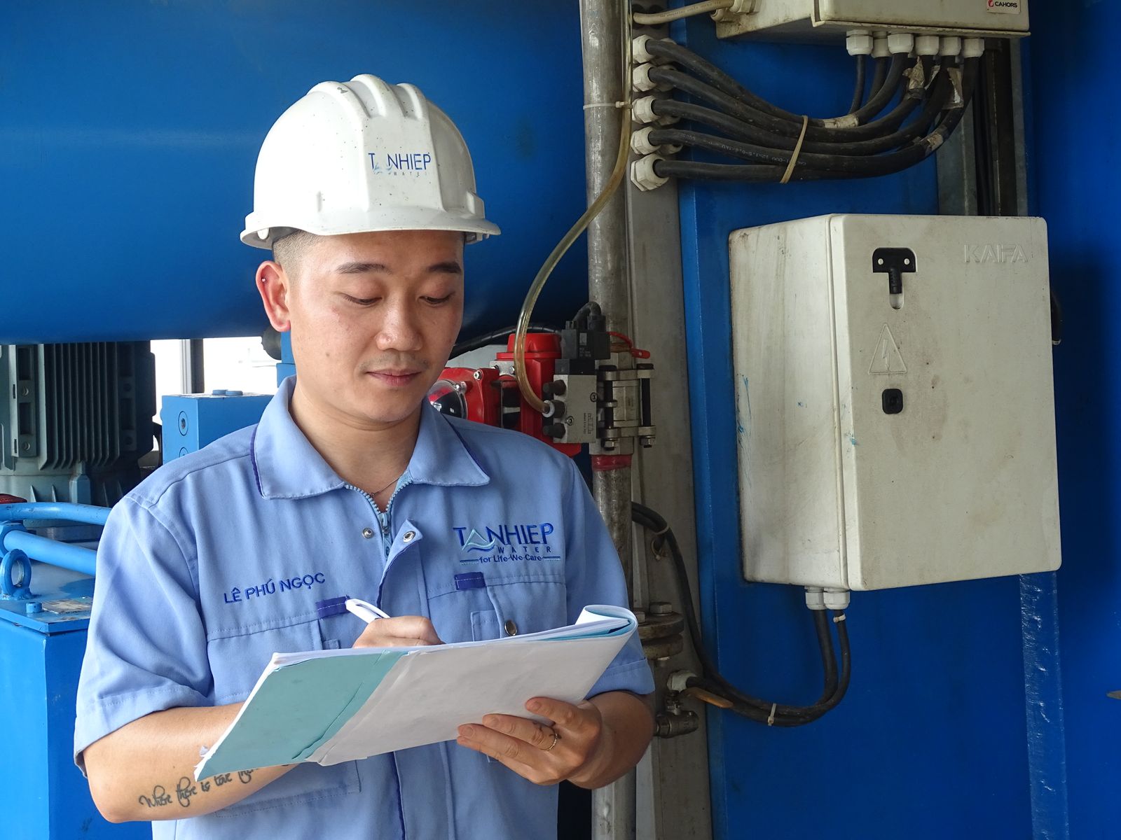 Técnico de capacete branco fazendo anotações em uma prancheta ao lado de um painel elétrico em uma planta industrial
