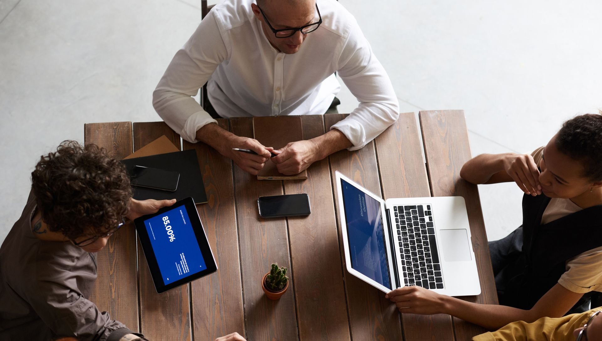 Vista de cima de uma equipe reunida em uma mesa de madeira, com laptop, tablet e celulares